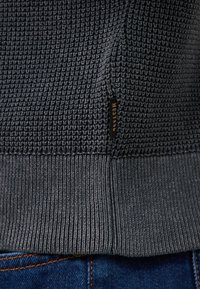 Saum eines dunkelgrauen Strickpullovers mit einem kleinen schwarzen Etikett mit der Aufschrift "MUSTANG", getragen über blauen Jeans.