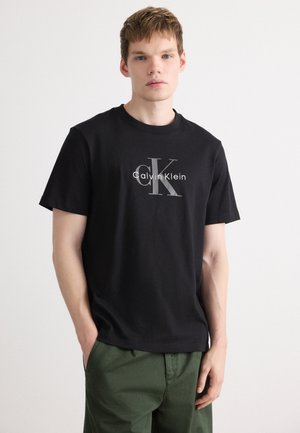 Mlad moški stoji z rokami v žepih, oblečen je v črno majico s tiskom Calvin Klein in zelene hlače na enobarvni podlagi.