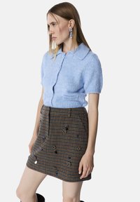 Lichtblauwe gebreide cardigan met korte mouwen en een kraag, gecombineerd met een bruine houndstooth rok met decoratieve versieringen.