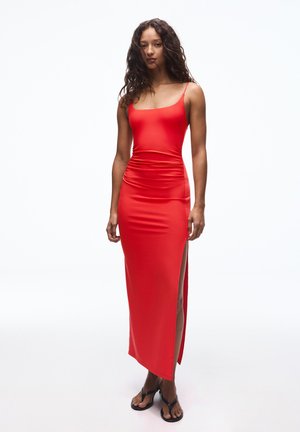 Žena oblečená v priliehavých červených šatách bez rukávov s rozparkom nad stehnom a v čiernych sandáloch, stojaca pred bielym pozadím.