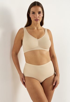 Vrouw met lang bruin haar die een beige draadloze BH en hooggetailleerd ondergoed draagt, staand tegen een witte muur, kijkend vooruit.