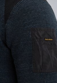 Niebieski sweter dzianinowy o strukturalnej powierzchni, z czarną łatą na rękawie, oznaczoną złotym napisem "PME LEGEND".