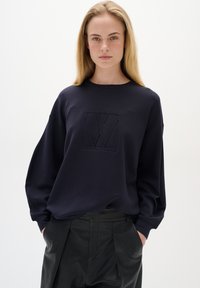 Tmavě modré mikina s texturovaným logem na přední straně, se zaobleným výstřihem a oversized rukávy, v kombinaci s černými kalhotami.