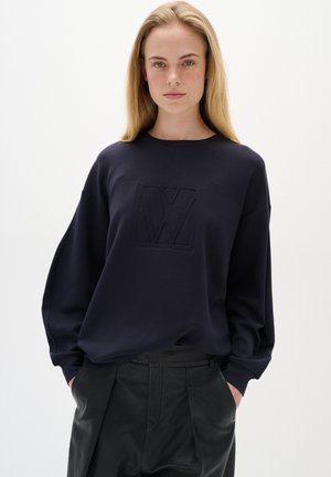Felpa blu navy con logo testurizzato sul davanti, caratterizzata da un collo rotondo e maniche oversized, abbinata a pantaloni neri.