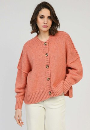 Femme aux cheveux bruns mi-longs portant un cardigan rose ample boutonné et un pantalon clair, debout devant un fond uni.