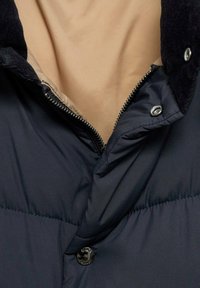 Marineblå puffjacka med dragkedja fram, knappdetaljer och mjukt beige innerfoder. Texturerad krage ger djup åt designen.