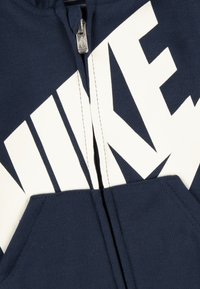 Navy blue zip-up hoodie made of cotton, featuring a large white "NIKE" logo, contrasting stitching, and a kangaroo pocket at the front.