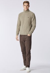 Maglione a collo alto beige in maglia treccia con motivo testurizzato, abbinato a pantaloni marroni e sneakers bianche. Design semplice e colori neutri.