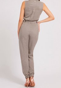 Žena v bezramenném taupe overal s tapered nohavicemi a elastickými manžetami, pózuje se rukou na boku, obutá v metalických růžově zlatých lodičkách.