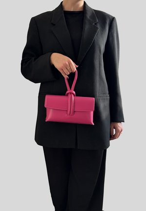 Persona che indossa un blazer e pantaloni neri, che tiene una piccola borsa in pelle rosa con manico ad anello, su uno sfondo neutro.