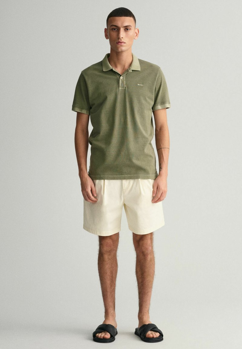 Polo vert avec col, manches courtes et logo brodé ; associé à un short beige clair et des sandales noires.