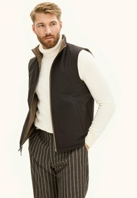 Brauner ärmelloser Weste mit hohem Kragen, Reißverschluss und Seitentaschen über einem cremefarbenen Zopfstrickpullover und gestreiften Hosen. Glattes, leichtes Material.