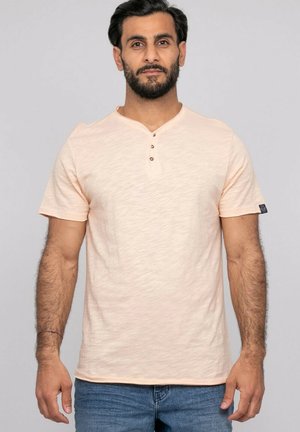 Homme aux cheveux foncés et à la barbe, portant un t-shirt henley à manches courtes couleur pêche clair et un jean bleu, debout devant un fond uni.