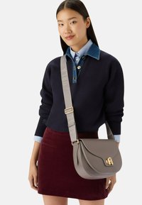 Femme portant un pull marine avec un col en denim et une jupe bordeaux, portant un sac en cuir taupe en bandoulière avec un fermoir doré et une large sangle grise.