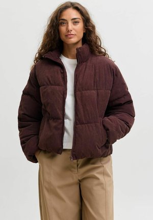 Woman with long curly hair wearing a dark brown corduroy puffer jacket over a white shirt and beige pants against a plain background.