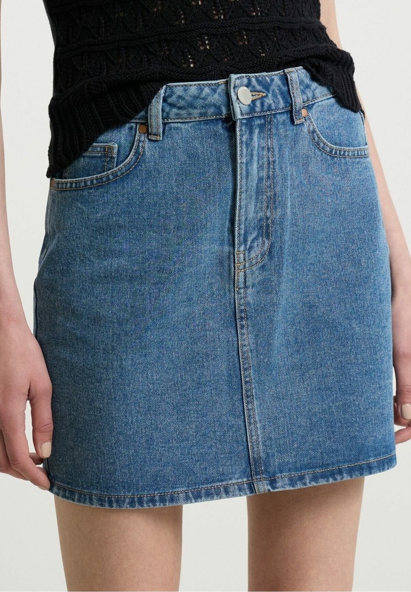 Femme portant une mini-jupe en denim bleu avec boutons sur le devant et poches, associée à un top en tricot noir texturé.