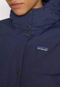 Navyblauwe Patagonia-jack met een hoge kraag, drukknoopsluiting en een gestikt quiltpatroon. Logo-patch op de linkerbovenborst.