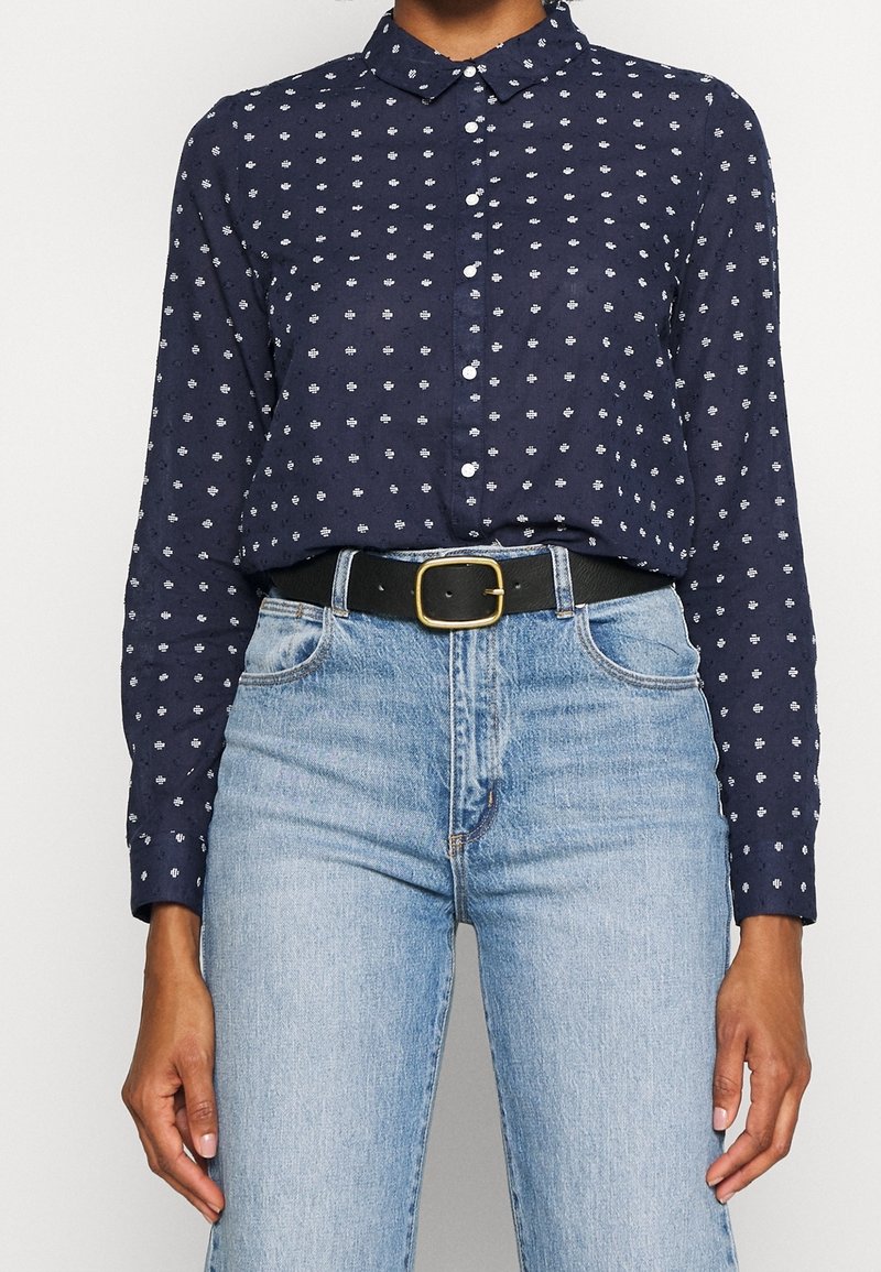 Personne portant une chemise bleu marine à boutons avec des motifs blancs, rentrée dans un jean bleu clair, et une ceinture noire avec une boucle dorée.