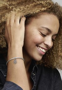 Strieborný náramok s textúrovaným dizajnom, ktorý obsahuje prívesok v tvare hamsa ruky s detailom oka, nosený na ruke s kučeravými vlasmi.