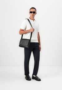 Schwarze Leder-Umhängetasche mit Klappenverschluss und verstellbarem Riemen, kombiniert mit einem enganliegenden weißen Poloshirt und dunklen Hosen.