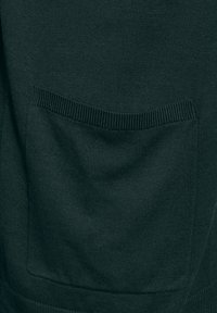 Mörkgrön stickad tyg har en ficka med ribbat detalj på framsidan, vilket visar en texturerad yta och en ren finish.