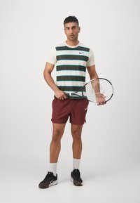 Hombre con una camisa a rayas y pantalones cortos rojos sosteniendo una raqueta de tenis, de pie contra un fondo claro y liso.