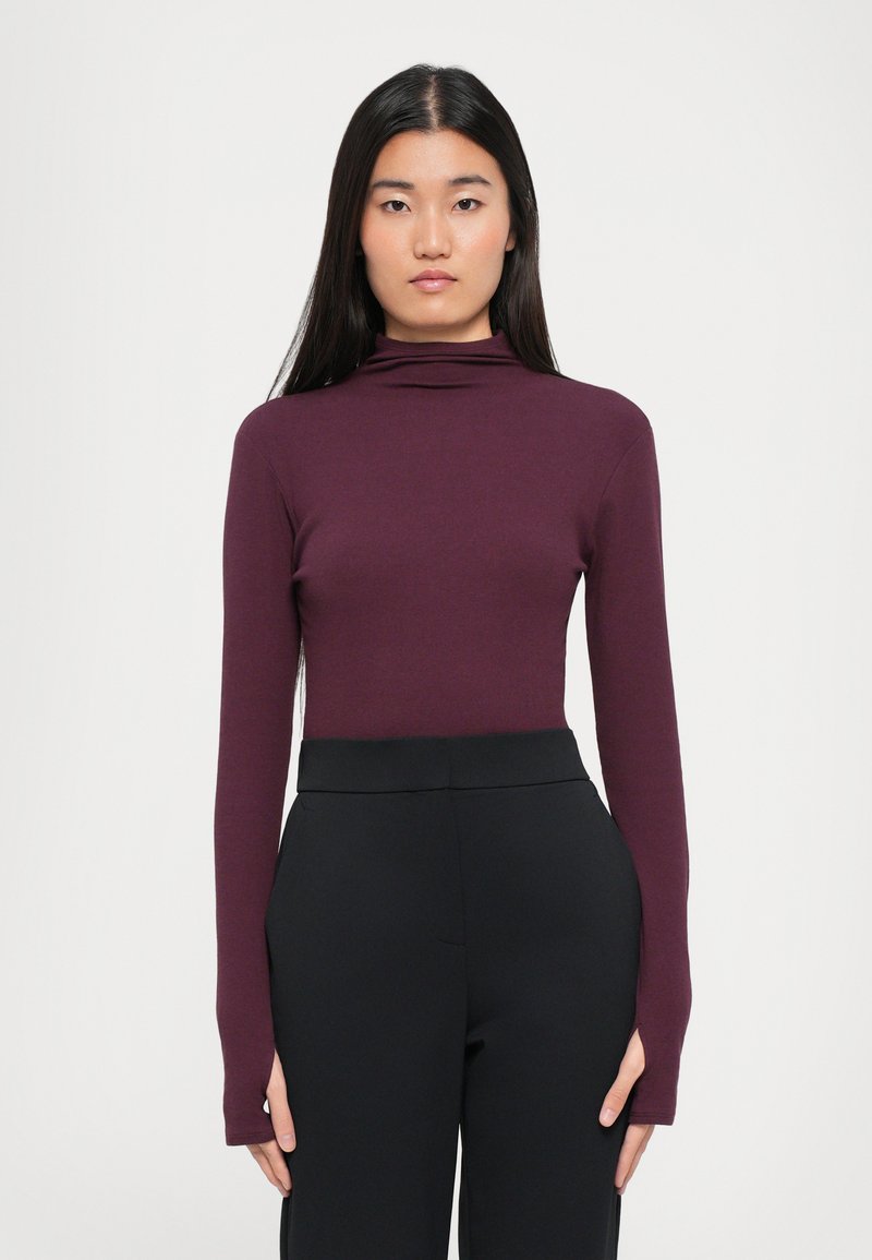 Long-sleeve, turtleneck top in deep burgundy fabric. Smooth texture, fitted design, paired with high-waisted black pants. Minimalist style.