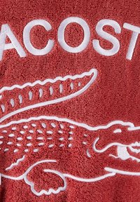 Serviette rouge en tissu coton texturé, avec le texte blanc brodé "LACOSTE" et un design détaillé de crocodile.