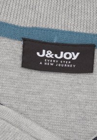 Zwart kledinglabel genaaid op grijs gebreid katoen met blauwe rand, met het merk "J&JOY" en de slogan "Elke stap een nieuwe reis."