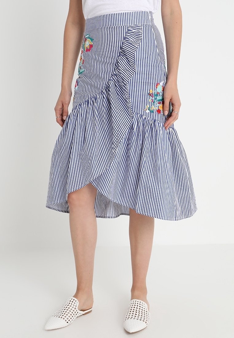 Jupe portefeuille à rayures bleues et blanches avec volants et broderies florales, portée avec des chaussures blanches à enfiler perforées.