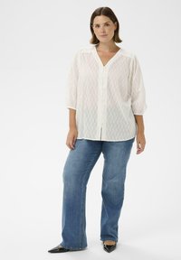 Witte blouse met knopen en een gemêleerd zigzagpatroon, driekwartmouwen, gecombineerd met blauwe flared jeans en zwarte puntige hakken.