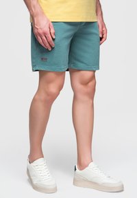 Mann mit türkisfarbenen Shorts, gelbem T-Shirt und weißen Turnschuhen steht vor einem schlichten hellen Hintergrund.