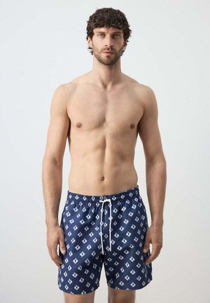 Man met donker haar en baard, gekleed in blauwe broek met patroon, staand tegen een effen lichte achtergrond.