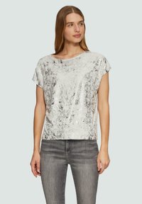 Srebrna bluzka z cekinami o luźnym kroju, szerokim dekolcie i krótkich rękawach, zestawiona z szarymi dżinsami. Gładka faktura z efektem połysku.