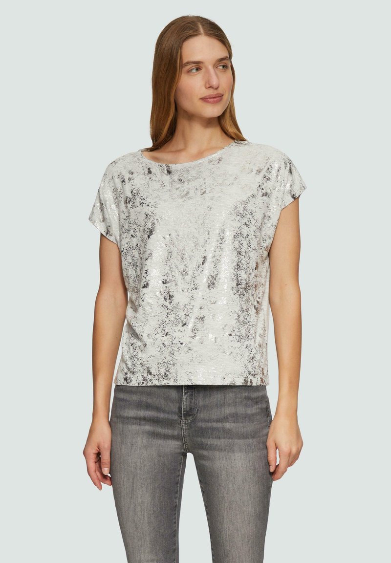 Srebrna bluzka z cekinami o luźnym kroju, szerokim dekolcie i krótkich rękawach, zestawiona z szarymi dżinsami. Gładka faktura z efektem połysku.