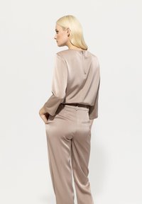 Beige satin two-piece outfit featuring a long-sleeve top with a back zipper and high-waisted trousers with side pockets. Smooth texture.
