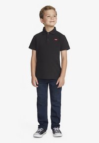 Svart poloshirt med to knapper og rød Levi's-logo; kombinert med mørkeblå jeans og svarte joggesko med hvite såler.