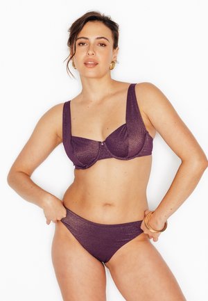 Donna che indossa un bikini viola glitterato con orecchini a cerchio dorati e braccialetti, posando contro uno sfondo bianco.