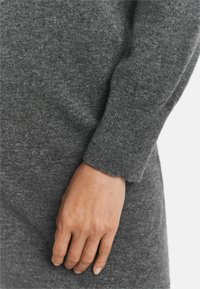 Maglione grigio in lana con un primo piano della manica e della mano. La texture sembra morbida, e il polsino è a coste per una vestibilità aderente.