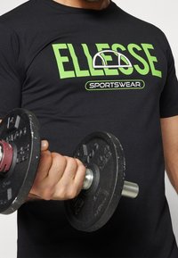 Černé tričko s krátkým rukávem s výrazným zeleným logem "ELLESSE" a textem "SPORTSWEAR". Osoba drží černou jednoruční činku.