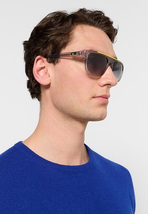 Uomo che indossa occhiali da sole con montatura trasparente con dettagli gialli e un maglione blu, rivolto verso destra su uno sfondo chiaro e semplice.