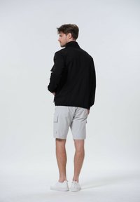 Schwarze Jacke mit hohem Kragen und langen Ärmeln, kombiniert mit hellgrauen Cargo-Shorts. Modell trägt weiße Sneakers. Glatte Stoffstruktur.