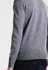 Grau gestrickter Pullover mit strukturiertem Muster, Rippbündchen und Saum sowie Rundhalsausschnitt, getragen über dunklen Jeans.