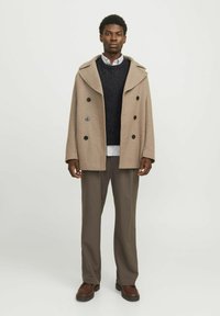 Abrigo beige de doble botonadura con un amplio cuello, botones negros, superpuesto a un suéter oscuro, combinado con pantalones topo y zapatos marrones.