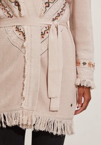 Beige knitted cardigan with intricate embroidery, fringed hem, and belt. Features colorful patterns along the edges and textured fabric.