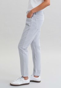 Světlé šedé slim-fit kalhoty vyrobené z bavlněné směsi, s bočními kapsami a jemnou texturou. V kombinaci s bílými teniskami.