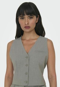 Grå, ærmeløs top med V-udskæring, der har knaplukning foran og en enkelt brystlomme. Lavet af tekstiler med struktur og en skræddersyet pasform.