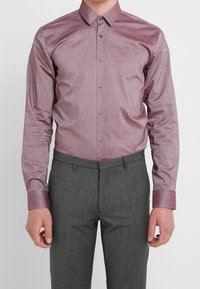 Camisa de manga larga color lila claro con botones, confeccionada en tela lisa, con cuello clásico y un sutil brillo, combinada con pantalones grises texturizados.