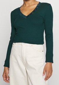 Tummanvihreä viistokaula-aukollinen pitkähihaisuus paita, jossa on musta pitsireunus kauluksessa. Yhdistetty vaaleankermanvärisiin korkeavyötäröisiin housuihin.