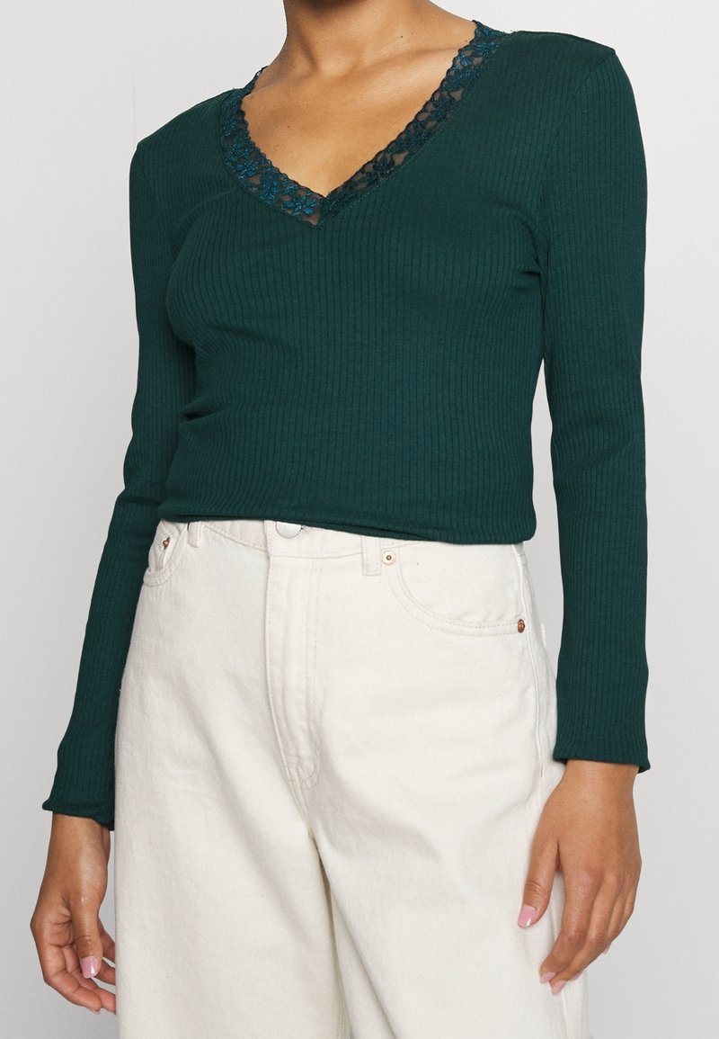 Tummanvihreä viistokaula-aukollinen pitkähihaisuus paita, jossa on musta pitsireunus kauluksessa. Yhdistetty vaaleankermanvärisiin korkeavyötäröisiin housuihin.